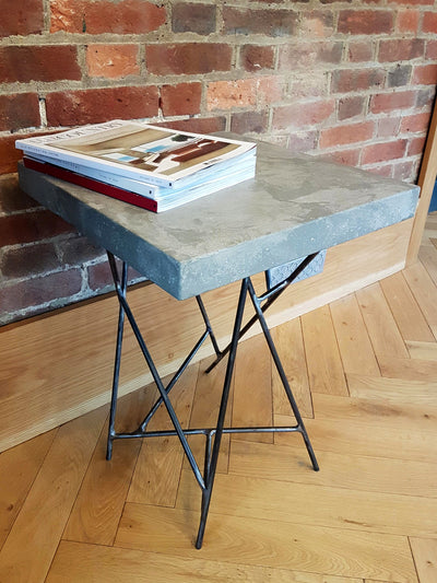 GFRC Concrete Side Tables with books on top, against a wall, with welded steel lattice legs. Prices starting from £750 each.  This client needed a pair of tables to position their ambient lighting. They chose to have random tones of natural shade concrete with movement through the concrete surface to reflect the colours of their existing concrete fireplace. The height of the Concrete tables and the size of the concrete table top were made to work with the height and size of the lamps and the fire hearth.